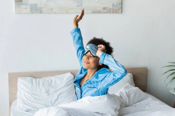 Sleepy african american woman in pajamas adjusting sleeping mask and stretching while smiling in bedroom — Stock Photo
