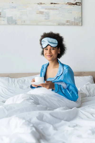 Угодила африканской американке в пижаме и маске для сна, держа чашку кофе в постели — стоковое фото