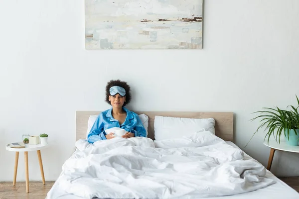 Femme afro-américaine gaie en pyjama et masque de sommeil tenant tasse de café au lit — Photo de stock