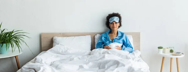 Mulher americana africana satisfeito em pijama azul e máscara de dormir segurando xícara de café na cama, banner — Fotografia de Stock