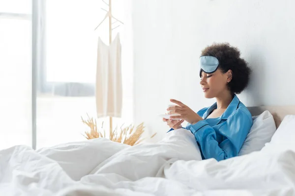 Joyful african american woman in pajamas and sleeping mask holding cup of coffee in bed — Stock Photo