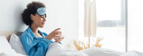 Mujer afroamericana complacida en pijama y máscara para dormir sosteniendo taza de café en la cama, pancarta - foto de stock