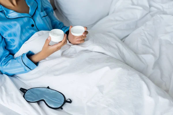 Vue recadrée d'une femme afro-américaine en pyjama tenant un récipient avec crème cosmétique au lit — Photo de stock