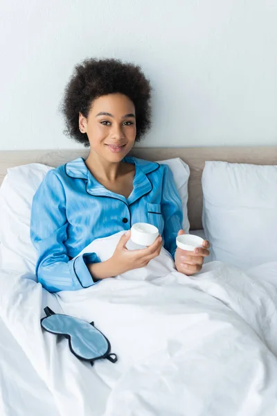 Mulher americana africana alegre em pijama segurando recipiente com creme cosmético na cama — Fotografia de Stock