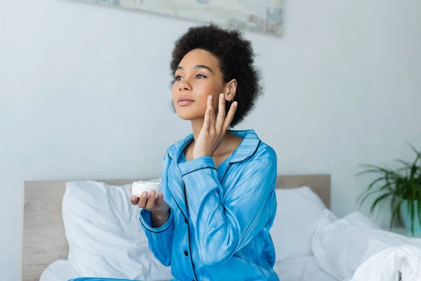 Femme afro-américaine bouclée en pyjama tenant le récipient et appliquant la crème visage dans la chambre — Photo de stock