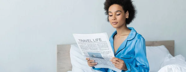 African american woman in pajamas reading travel life newspaper in bedroom, banner — Stock Photo