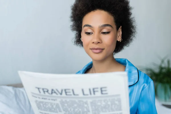 Mujer afroamericana en pijama leyendo borrosa vida de viaje periódico en el dormitorio - foto de stock