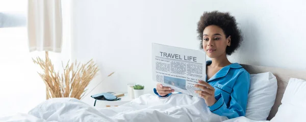 Joven afroamericana mujer en pijama leyendo viaje vida periódico en cama, pancarta - foto de stock