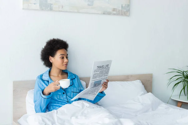 Mujer afroamericana en pijama leyendo el periódico de viaje mientras sostiene una taza de café en el dormitorio - foto de stock