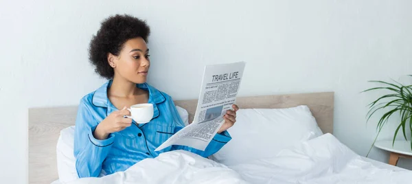 African american woman in pajamas reading travel life newspaper while holding cup of coffee in bedroom, banner — Stock Photo