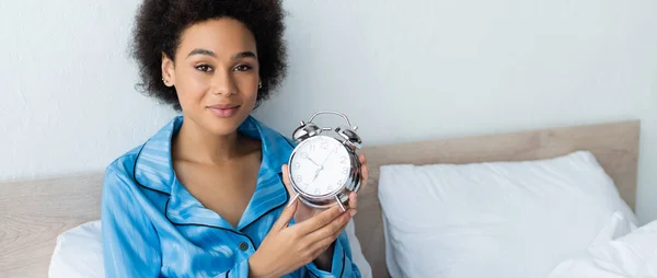 Smiling african american woman in pajamas holding alarm clock in bedroom, banner — Stock Photo