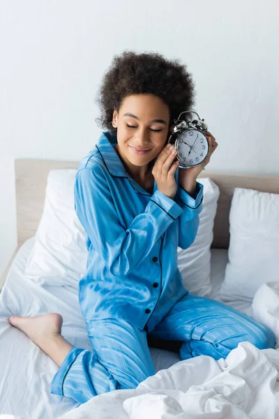 Smiling african american woman with closed eyes holding alarm clock in bedroom — Stock Photo