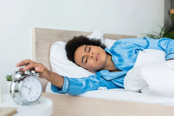 Sleepy african american woman in pajamas frowning while reaching alarm clock — Stock Photo