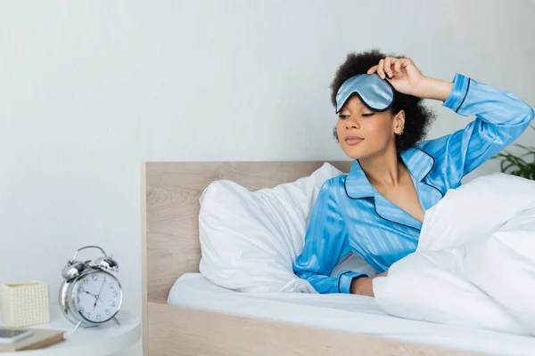 Mulher americana africana acordada em pijama ajustando máscara de dormir enquanto olha para o despertador no quarto — Fotografia de Stock