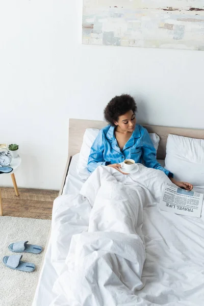 High angle view of african american woman in pajamas reading travel life newspaper while holding cup of coffee in bed — Stock Photo