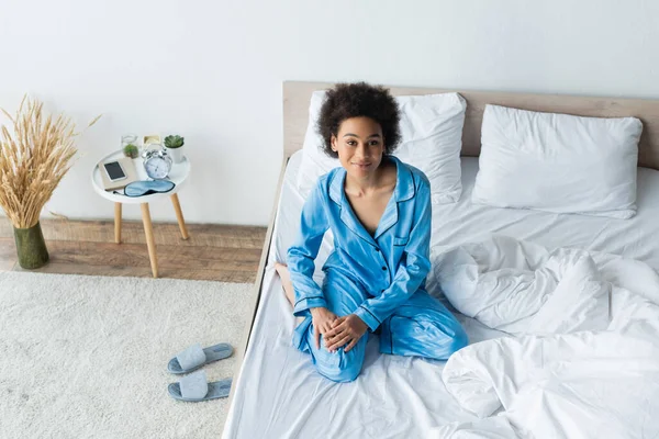 Vue grand angle de femme afro-américaine heureuse en pyjama regardant la caméra dans la chambre — Photo de stock