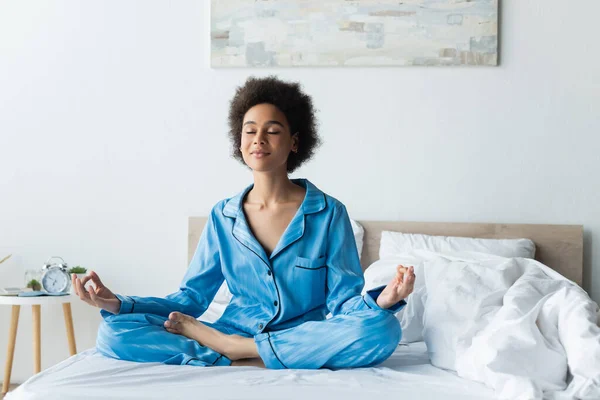 Mujer afroamericana complacida en pijama meditando en la cama - foto de stock