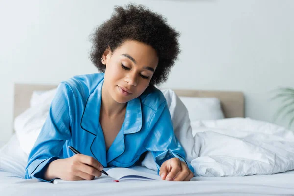 Glückliche afrikanisch-amerikanische Frau im Pyjama auf dem Bett liegend und auf Notizbuch schreibend — Stockfoto