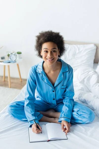 Mulher americana africana alegre em pijama sentado na cama e escrevendo no caderno — Fotografia de Stock