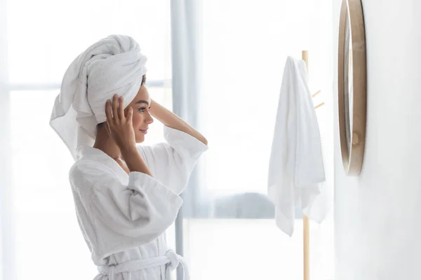 Alegre afroamericana mujer ajustando toalla y mirando espejo en cuarto de baño - foto de stock