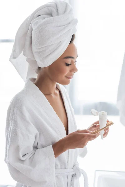 Femme afro-américaine en peignoir et serviette tenant dentifrice et brosse à dents dans la salle de bain — Photo de stock