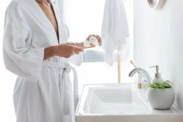 Vue recadrée de femme afro-américaine en peignoir tenant dentifrice et brosse à dents dans la salle de bain — Photo de stock