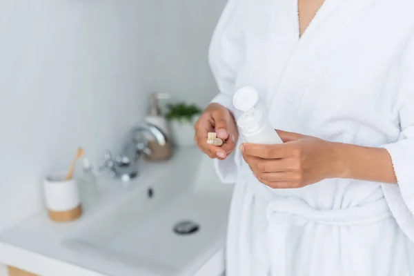 Vue recadrée de femme afro-américaine en peignoir tenant tube avec dentifrice et brosse à dents dans la salle de bain — Stock Photo