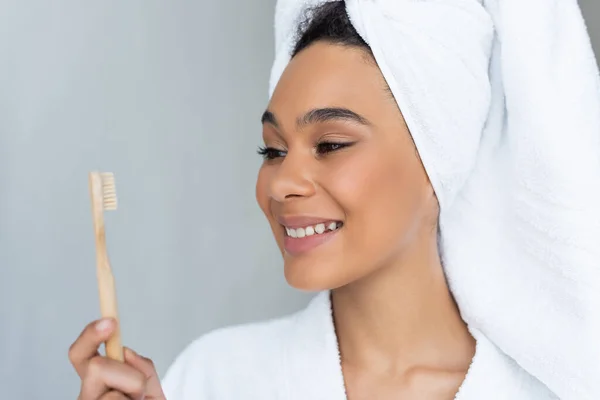 Heureuse femme afro-américaine en peignoir et serviette tenant brosse à dents dans la salle de bain — Photo de stock