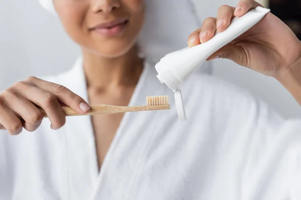 Vista recortada de mujer afroamericana borrosa en albornoz sosteniendo pasta de dientes y cepillo de dientes en el baño - foto de stock