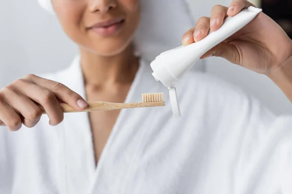 Vue recadrée de femme afro-américaine floue en peignoir tenant dentifrice et brosse à dents dans la salle de bain — Photo de stock