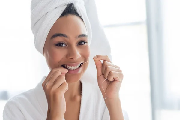 Sorrindo jovem afro-americana mulher em roupão de banho dentes de fio dental no banheiro — Fotografia de Stock