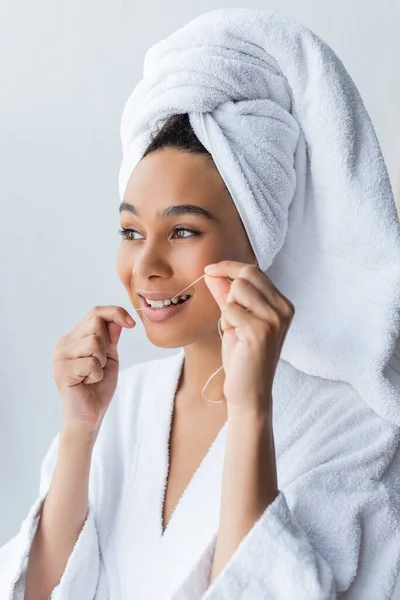 Alegre joven afroamericana mujer en albornoz hilo dental en baño - foto de stock