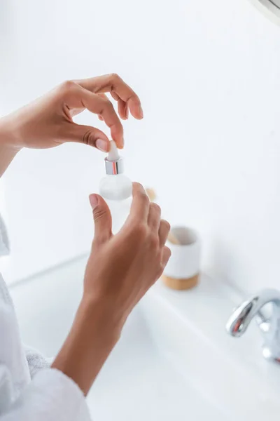 Cropped view of young african american woman holding bottle with serum in bathroom — Stock Photo