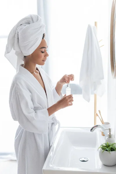 Side view of young african american woman in white bathrobe holding bottle with serum in bathroom — Stock Photo