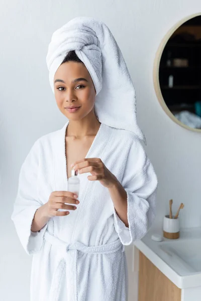 Jeune femme afro-américaine en serviette blanche et peignoir tenant bouteille avec sérum dans la salle de bain — Photo de stock