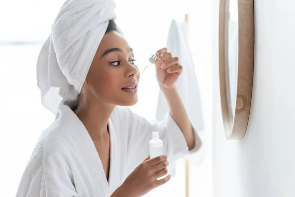 Молода афроамериканка в білій халаті, тримаючи пляшку в руках і прикладаючи сироватку на обличчі біля дзеркала. — стокове фото