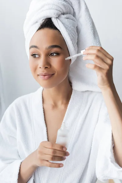Erfreut afrikanisch-amerikanische Frau im weißen Bademantel hält Flasche und Anwendung von Serum im Badezimmer — Stockfoto
