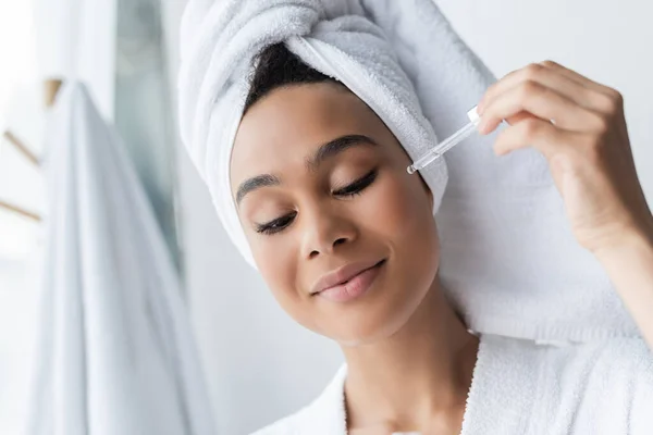 Heureux afro-américaine en serviette blanche appliquer sérum dans la salle de bain — Photo de stock