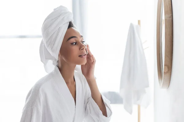 Young african american woman in towel cleansing face with cotton pad near mirror — Stock Photo