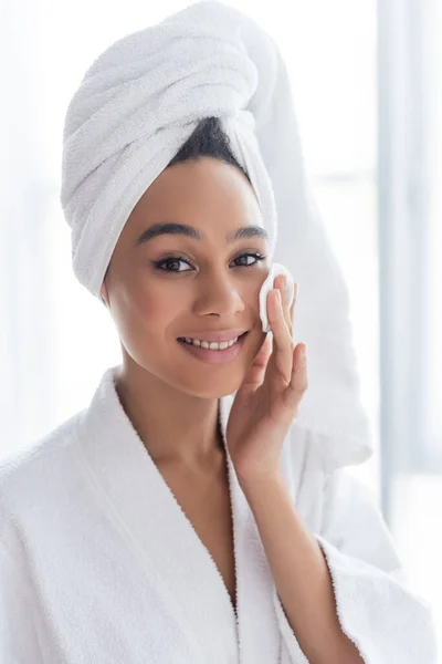 Fröhliche afrikanisch-amerikanische Frau in Handtuch Reinigung Gesicht mit Baumwollkissen im Badezimmer — Stockfoto