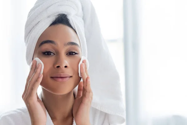 Giovane donna afroamericana in tovagliolo viso detergente con batuffoli di cotone in bagno — Foto stock