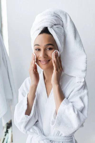 Alegre africana americana mujer en toalla limpieza cara con almohadillas de algodón en baño - foto de stock