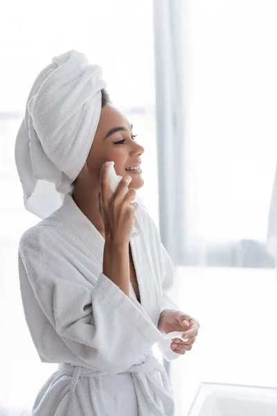 Heureuse femme afro-américaine en serviette tenant bouteille avec nettoyant et coton tampon dans la salle de bain — Photo de stock