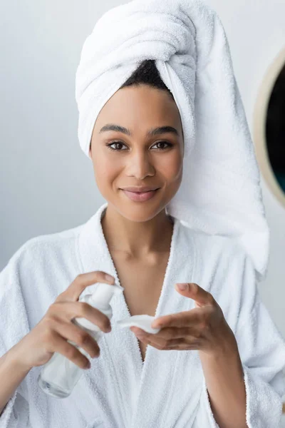 Mujer americana africana alegre en botella de la celebración de la toalla con el limpiador y la almohadilla de algodón en cuarto de baño - foto de stock