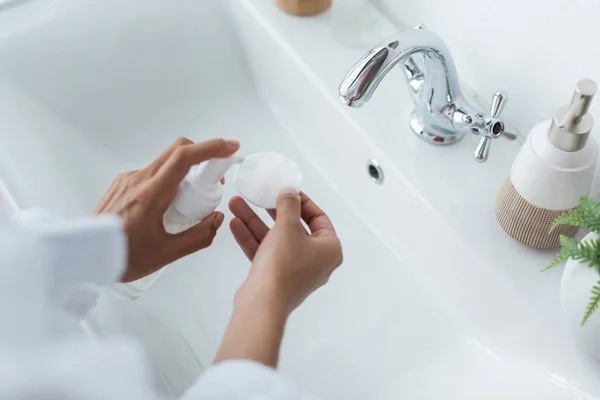 Abgeschnittene Ansicht einer afrikanisch-amerikanischen Frau, die eine Flasche mit Reinigungsmittel und Baumwollkissen in der Nähe des Waschbeckens hält — Stockfoto