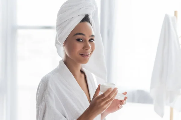 Femme afro-américaine souriante en peignoir contenant avec crème visage dans la salle de bain — Photo de stock