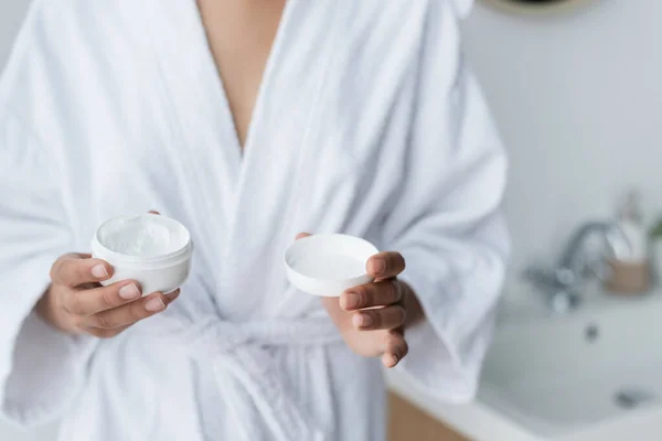 Vue partielle de la femme afro-américaine en peignoir contenant avec crème visage dans la salle de bain — Photo de stock