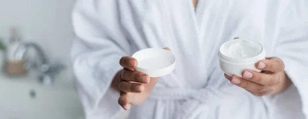 Vue partielle de la femme afro-américaine en peignoir contenant avec crème visage dans la bannière de salle de bains — Photo de stock