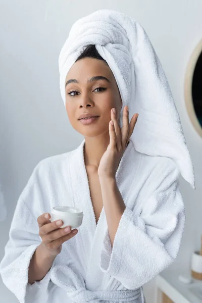 Mulher afro-americana em roupão segurando recipiente e aplicando creme facial no banheiro — Fotografia de Stock