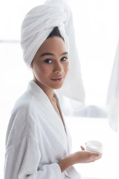 Mulher africana americana em roupão de banho segurando recipiente com creme cosmético no banheiro — Fotografia de Stock